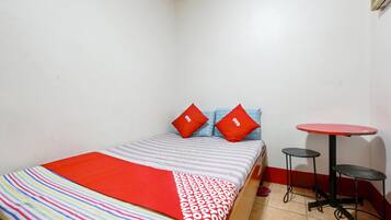 A small, neatly arranged room with a bed, two pillows, a striped blanket, and a small round table with a stool.
