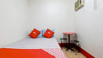 A small, narrow room with a bed, two red pillows, a striped blanket, a small round table with two stools, and a wall-mounted air vent.