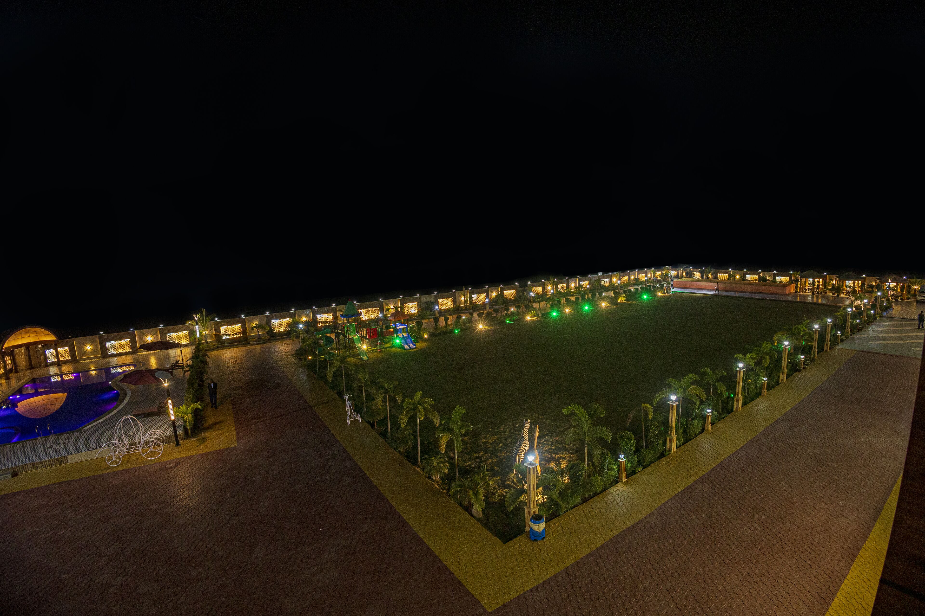 A large, well-lit outdoor area with a swimming pool, palm trees, and a pathway lined with lights.