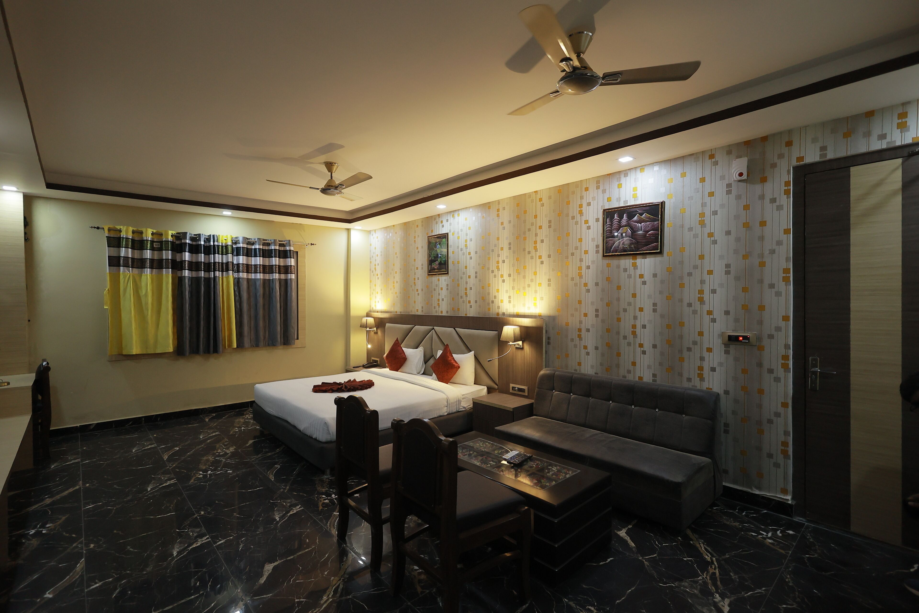 A hotel room with a bed, a sofa, a desk, and a chair. There are ceiling fans, a wall-mounted lamp, and a picture on the wall.