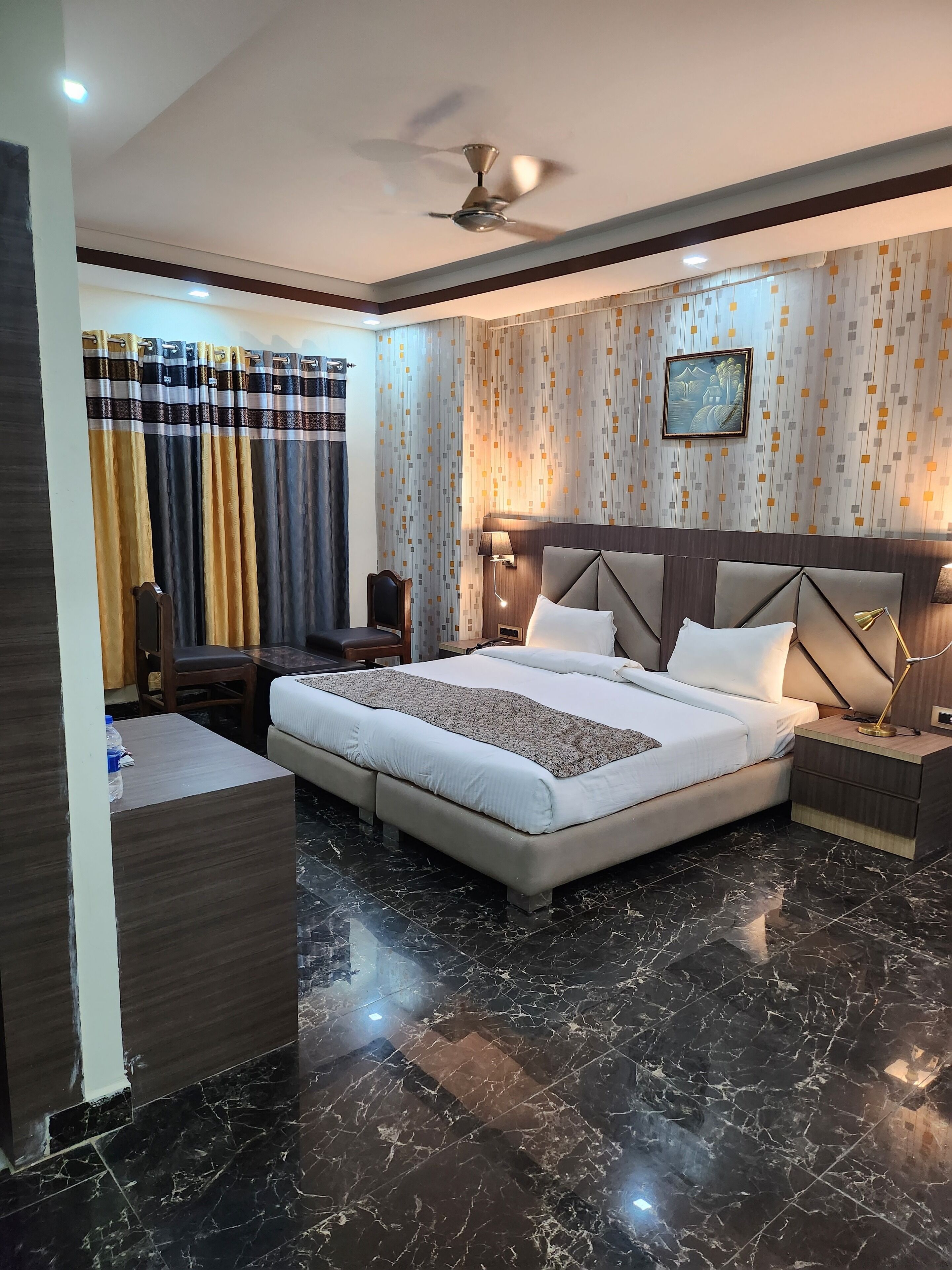 A modern hotel room with a large bed, a desk, a chair, a ceiling fan, and a wall-mounted lamp.