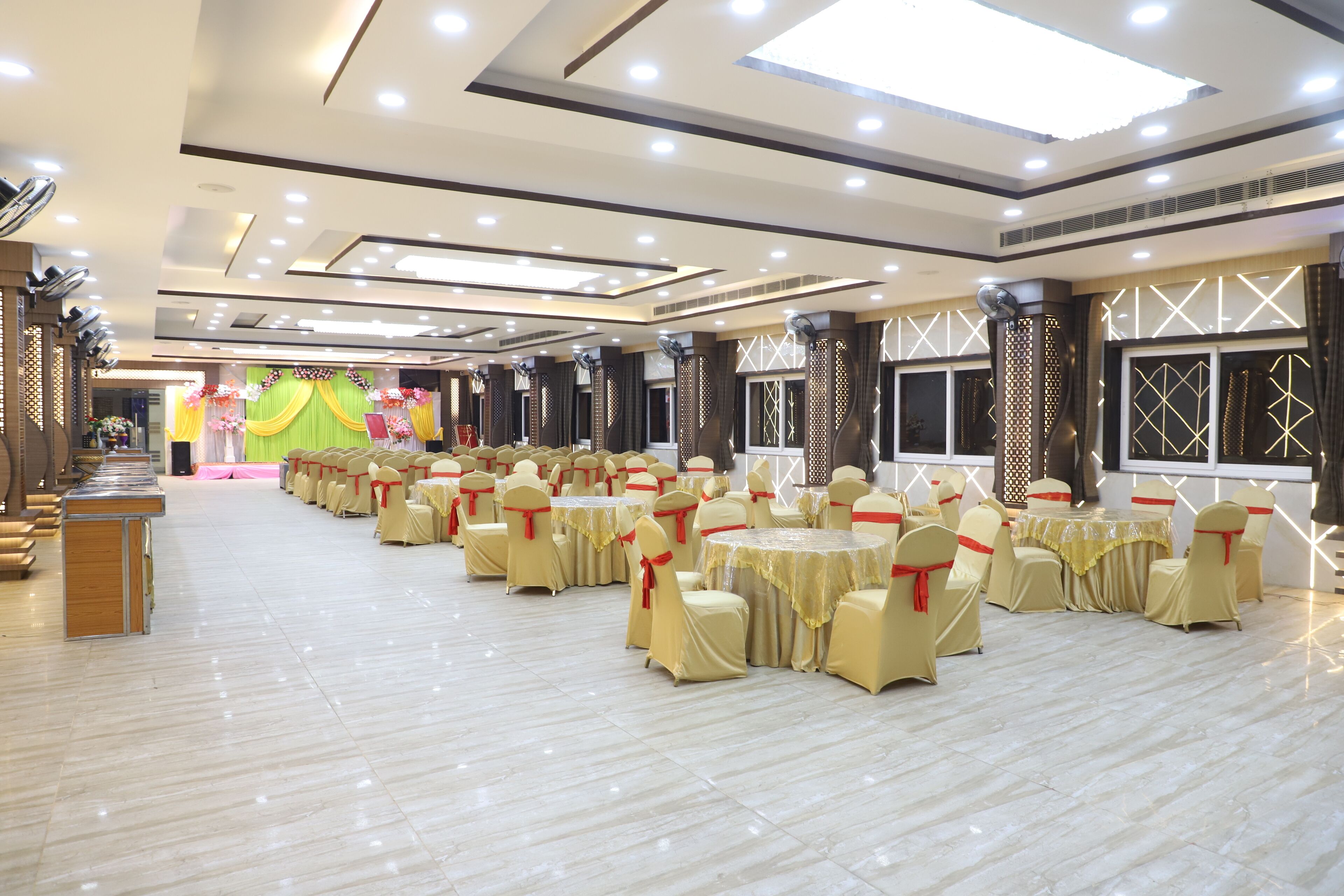 Un amplio salón de banquetes con filas de mesas y sillas redondas, un escenario con adornos florales y un diseño de techo moderno.