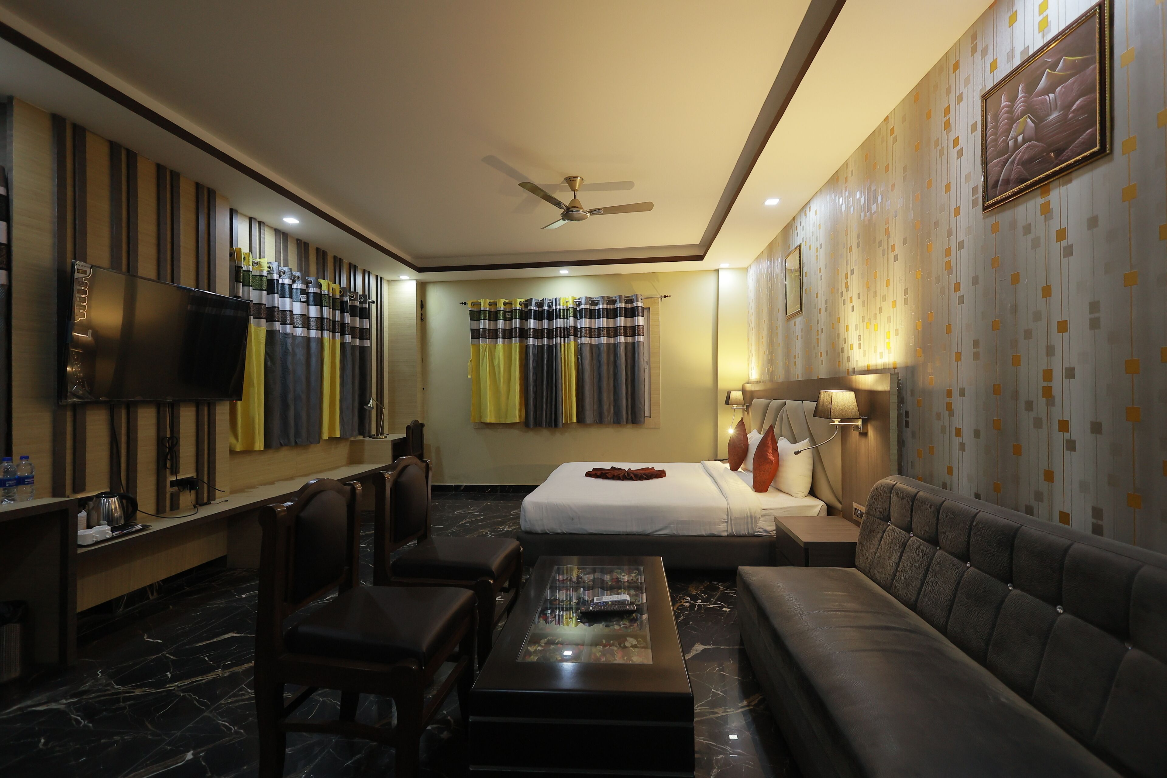 A modern hotel room with a flat-screen TV, a sofa, a coffee table, a bed with a decorative headboard, a ceiling fan, and a patterned wall.