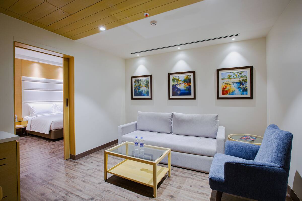 A hotel room with a bed, a sofa, a blue armchair, a glass coffee table, and three framed pictures on the wall.