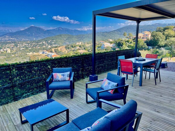 A wooden deck with outdoor furniture, including chairs and a table, overlooking a scenic view of a mountainous landscape.
