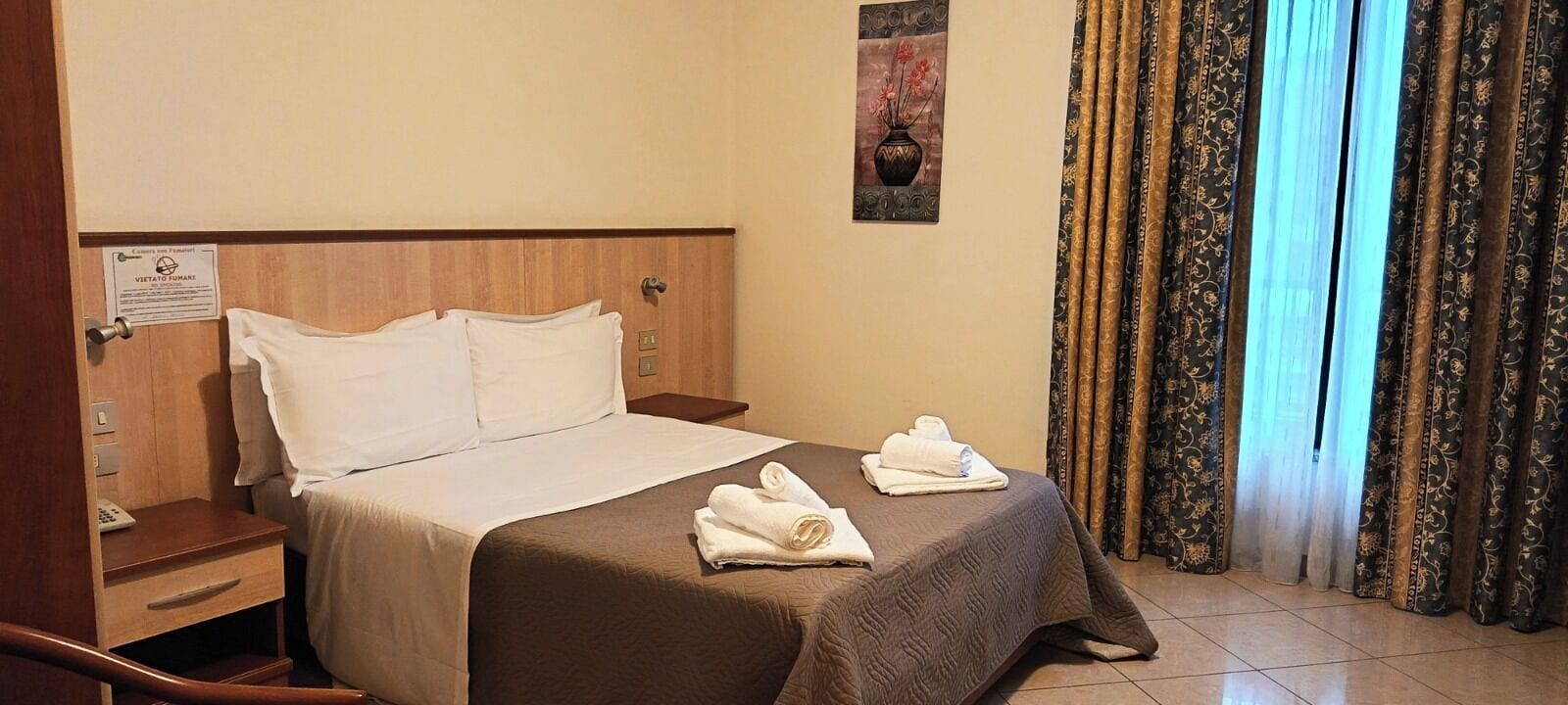 Camera d'albergo con un letto, due cuscini, un comodino con il telefono, una targhetta a parete e una finestra con tende.