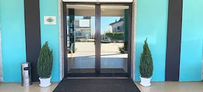 Entrada de hotel con puerta de vidrio, plantas en macetas y un basurero.