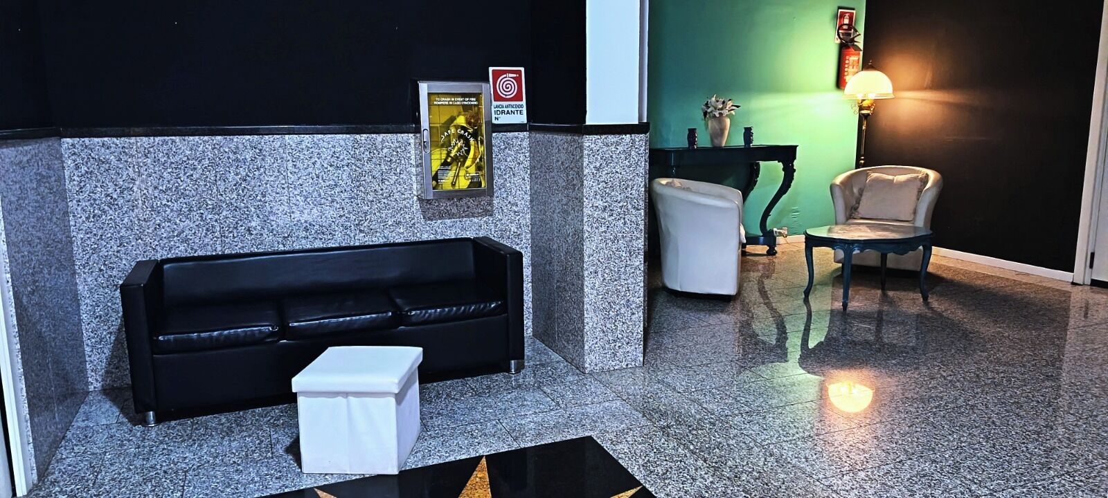 A lobby with a black leather sofa, a white side table, a small white trash bin, a black and white wall with a poster, a green wall with a lamp, and a chair.