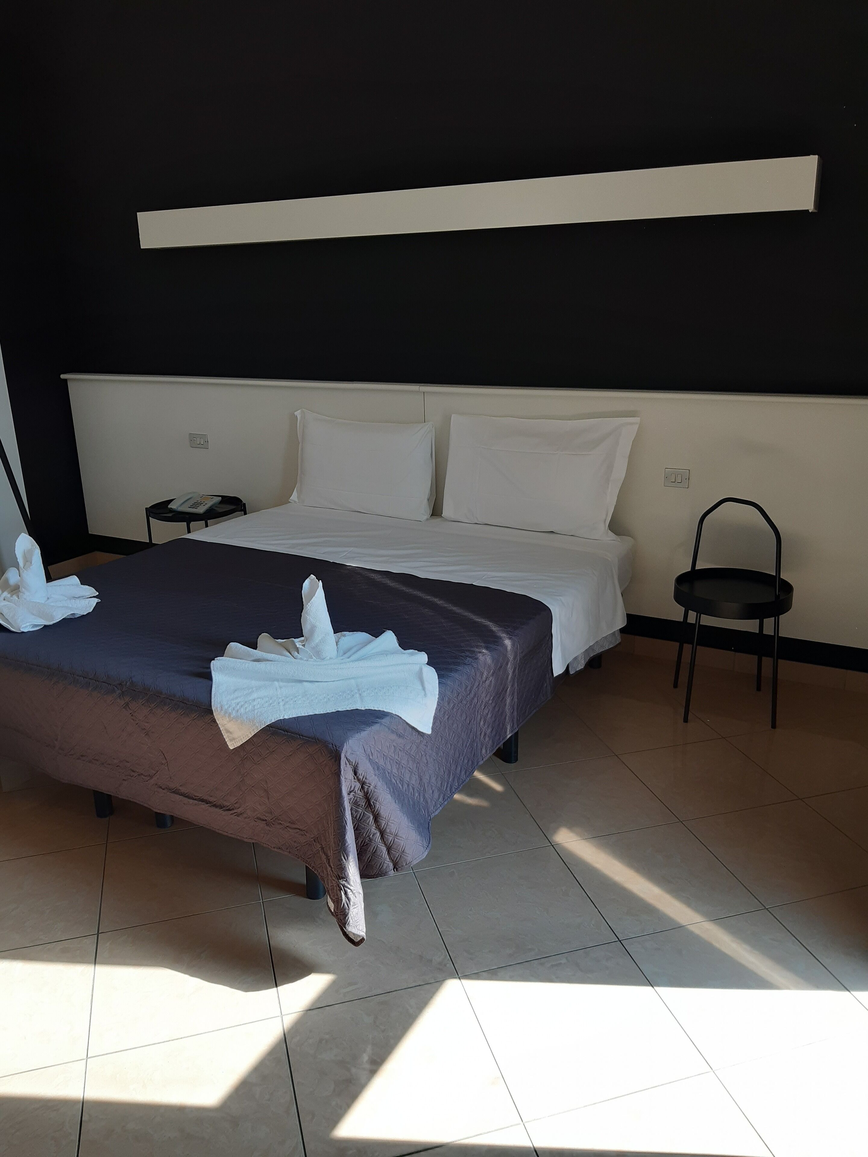 A neatly made bed with a dark-colored bedspread and white pillows, a bedside table with a phone, and a chair in a room with tiled flooring and a black wall.