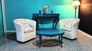 Une pièce avec deux fauteuils blancs, une table bleue, une petite lampe de table bleue et un vase rempli de fleurs, posé sur une petite table devant un mur de couleur turquoise.