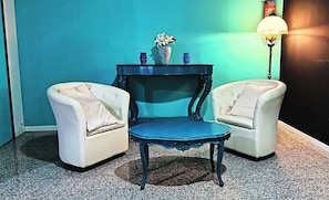 Una habitación con dos sillones blancos, una mesa azul, una pequeña lámpara de mesa azul y un jarrón con flores sobre una mesa pequeña frente a una pared verde azulado.