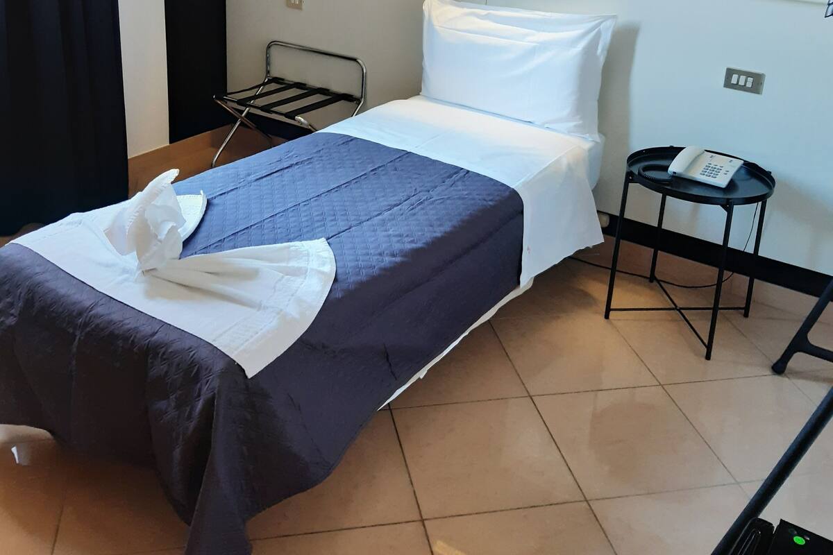Ein ordentlich bezogenes Bett mit einer blau-weißen Tagesdecke, einem weißen Kopfkissen und einem Nachttisch mit einem Telefon.