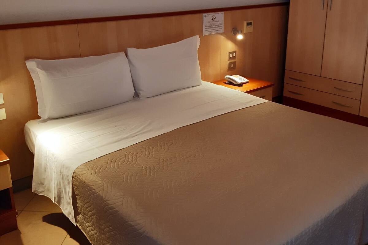 Ein ordentlich bezogenes Bett mit weißen Leinen, ein Holz-Kopfteil, ein Nachttisch mit Lampe und ein Kleiderschrank.