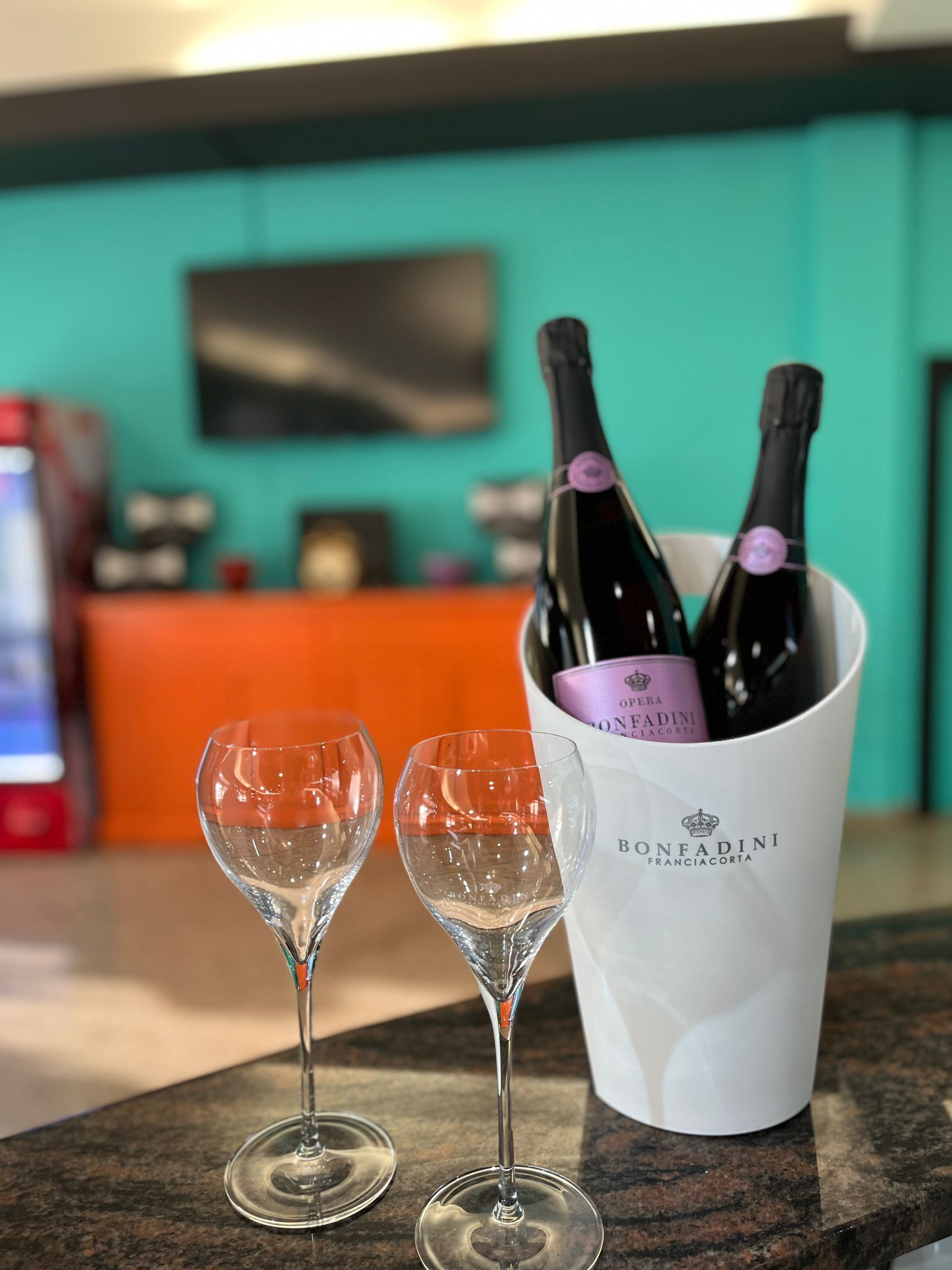 Dos copas de vino vacías sobre una encimera, un cubo blanco con dos botellas de vino, y una pared color esmeralda con un televisor de pantalla plana montado sobre la encimera.