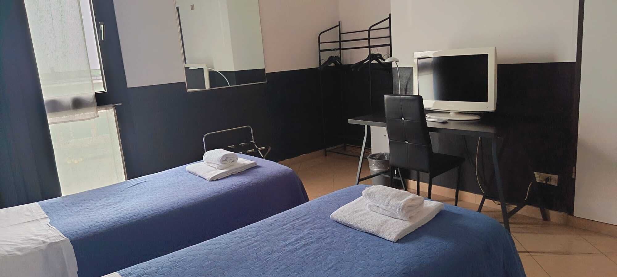 A hotel room with two beds, a desk with a computer and TV, and a window.