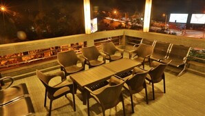 Eine Dachterrasse mit einem langen Tisch und Stühlen, einer kleinen Bar und Blick auf die beleuchtete Stadt.