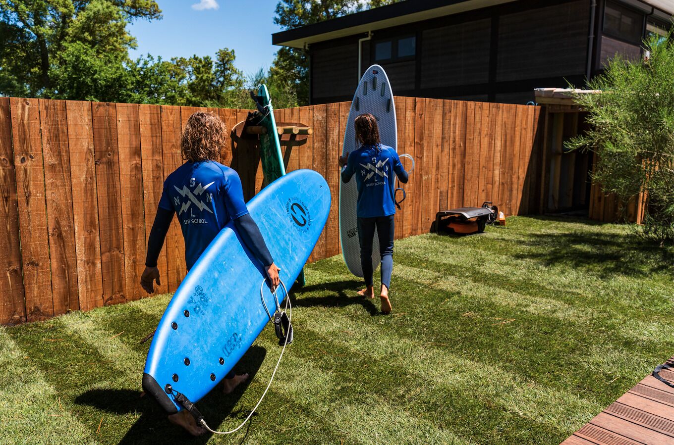 Two people in blue wetsuits carrying surfboards in a backyard with a wooden fence and a house in the background.