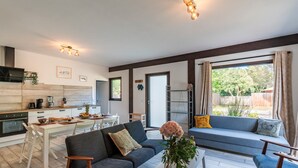 Ett modernt vardagsrum med en soffa, ett soffbord och en matplats. Det finns ett kök med en spis och ett matbord med stolar. Rummet har stora fönster och utsikt över omgivningen.