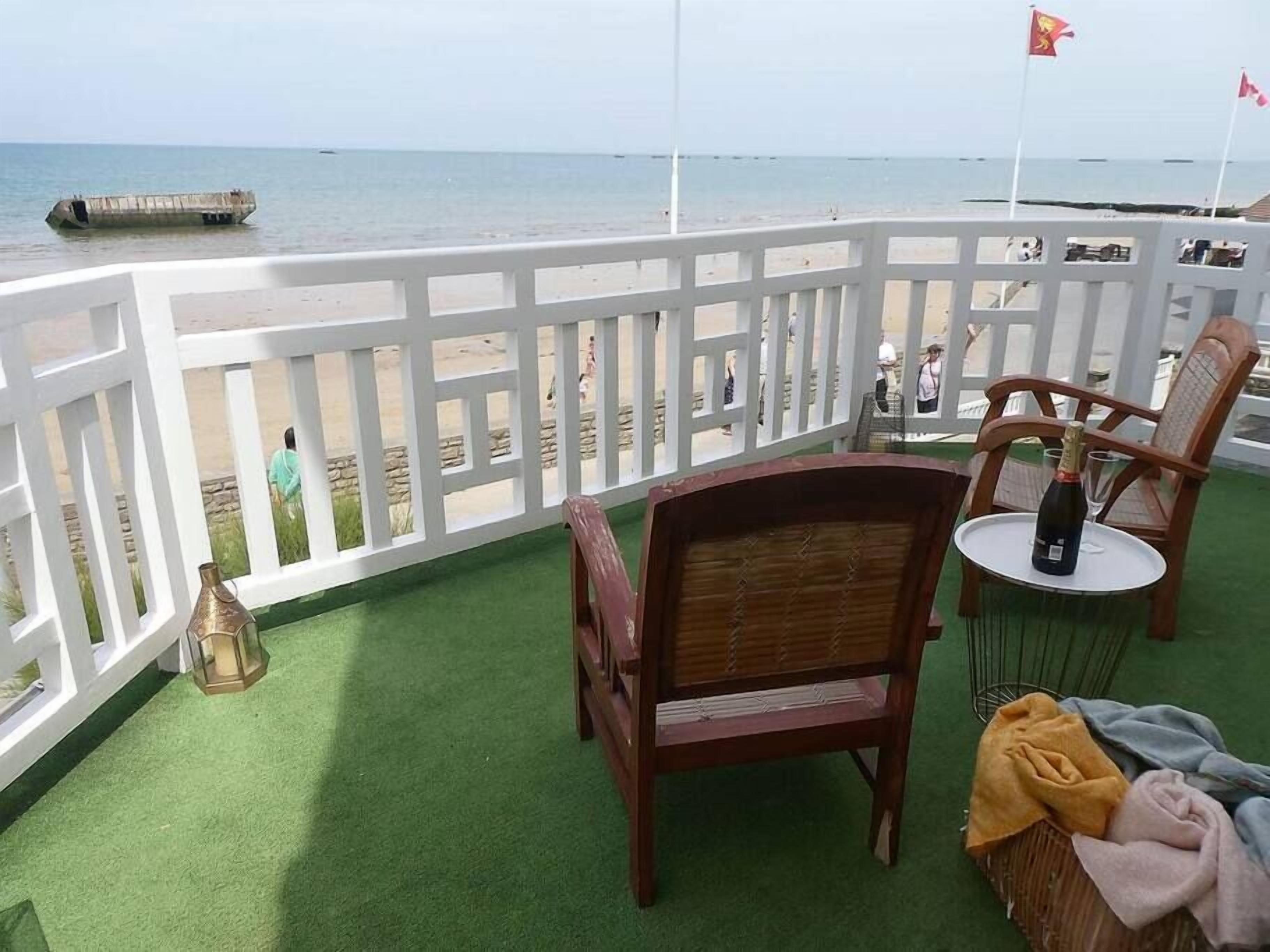 A balcony with a view of the beach, a wooden chair, a small table with a bottle, and a flag.