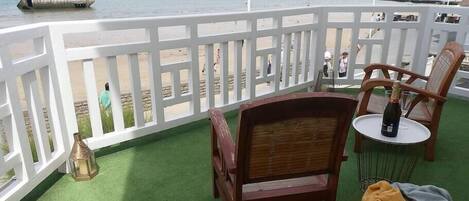 A balcony with a view of the beach, a wooden chair, a small table with a bottle, and a flag.