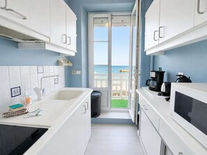 Une cuisine étroite avec des meubles blancs, une machine à café noire et une vue sur la mer par une porte vitrée.