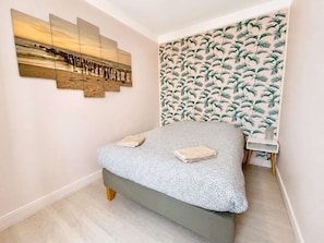 Un dormitorio con cama, una obra de arte en la pared, un papel pintado estampado y una mesita de noche pequeña.