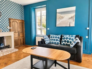 Una habitación con pared azul, sofá negro con cojines estampados, una mesa de centro de madera, chimenea y ventana con vistas al exterior.