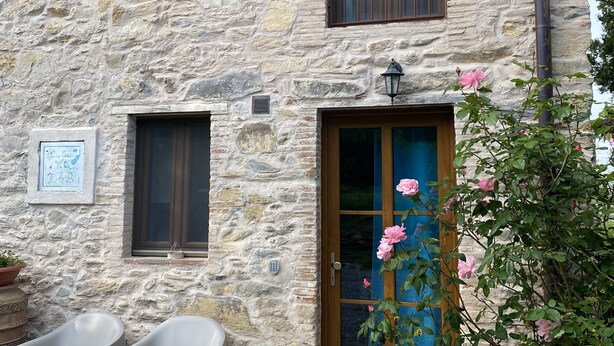 Un edificio de piedra con puerta y ventanas de madera, dos sillas de plástico afuera y una maceta con flores rosas.