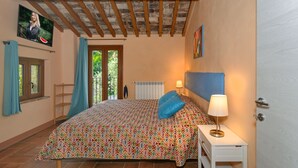 Un dormitorio con una colcha estampada, una mesita de noche con lámpara, un televisor en la pared y una ventana con cortinas.