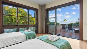 Quarto de hotel com duas camas, uma grande porta de vidro deslizante para área externa com cadeiras e uma mesa, e vista para árvores e céu.