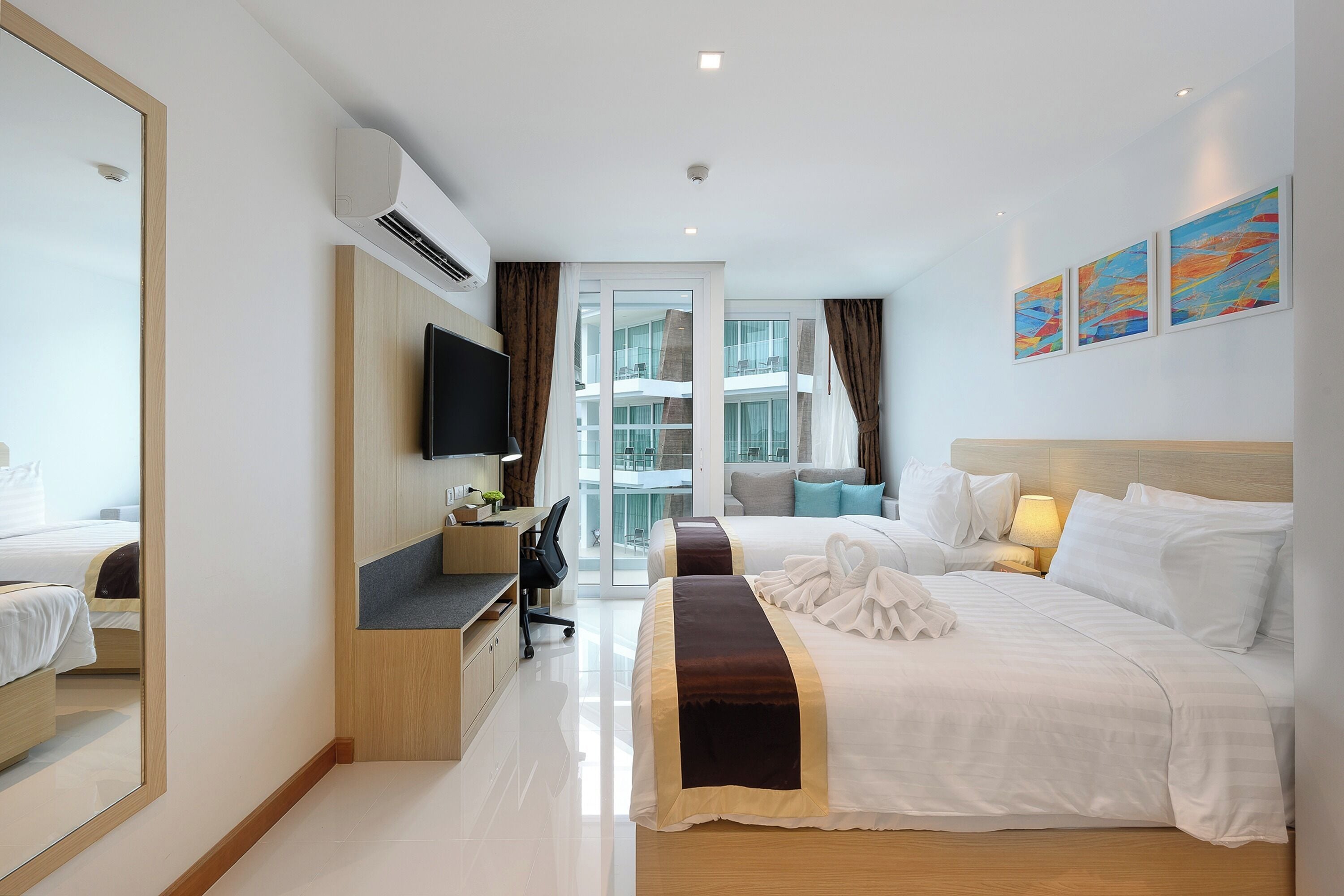 A hotel room with a large bed, a desk, a chair, a TV, and a balcony with a view of the building.
