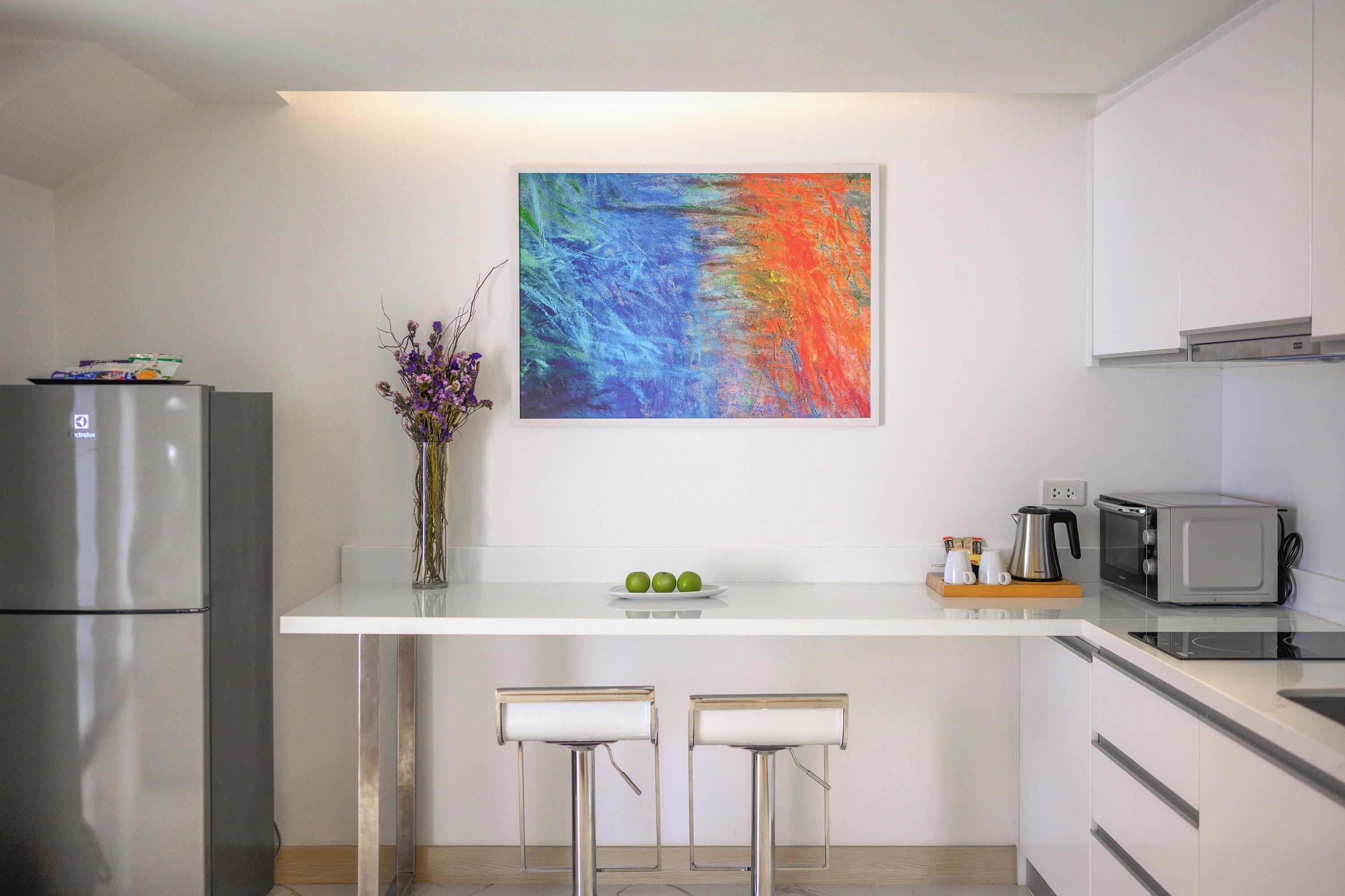 A modern kitchen with a white countertop, two bar stools, a refrigerator, a microwave, and a colorful abstract painting on the wall.