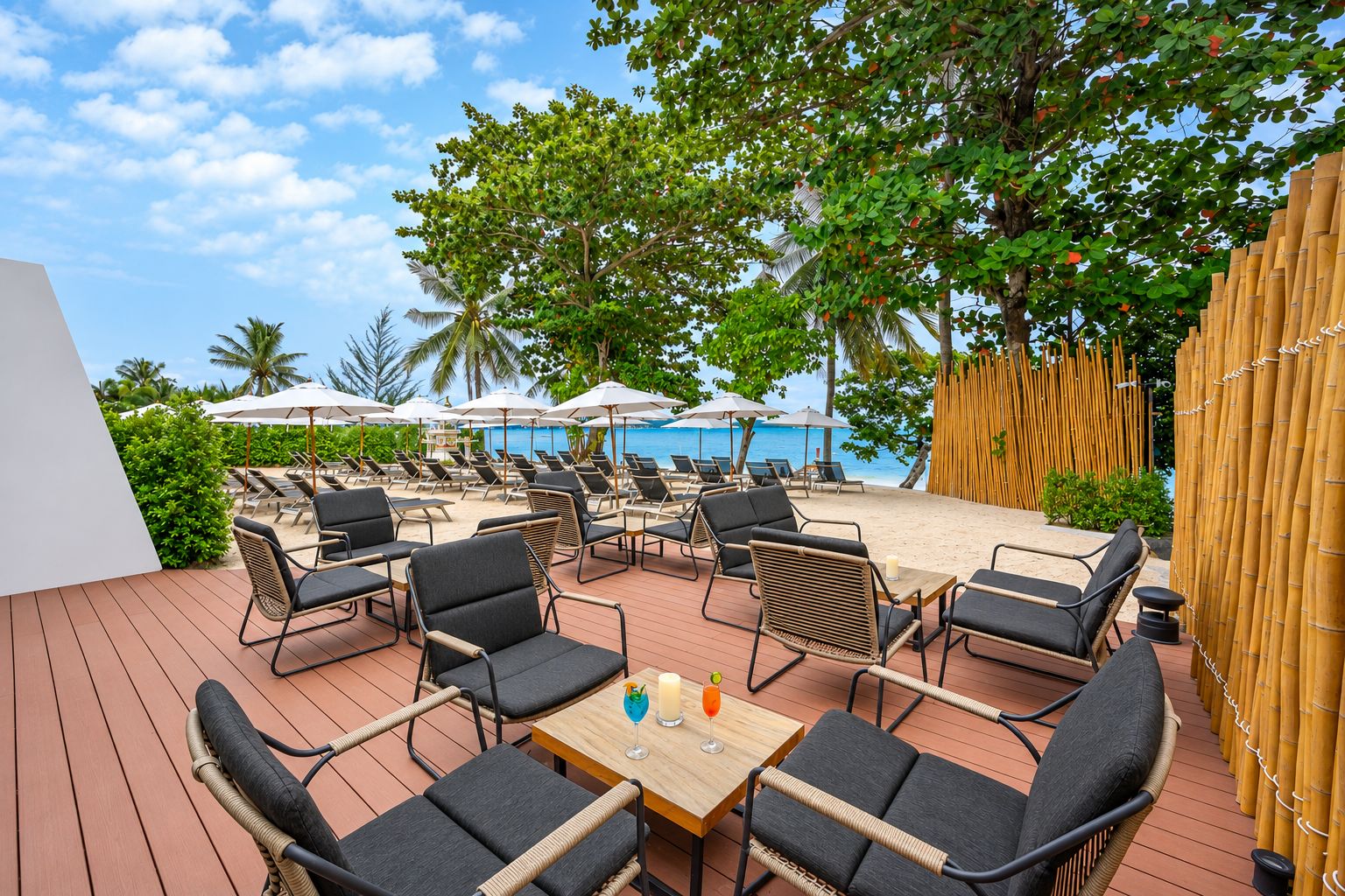 A deck with outdoor seating and umbrellas, overlooking a beach with palm trees and a clear sky.