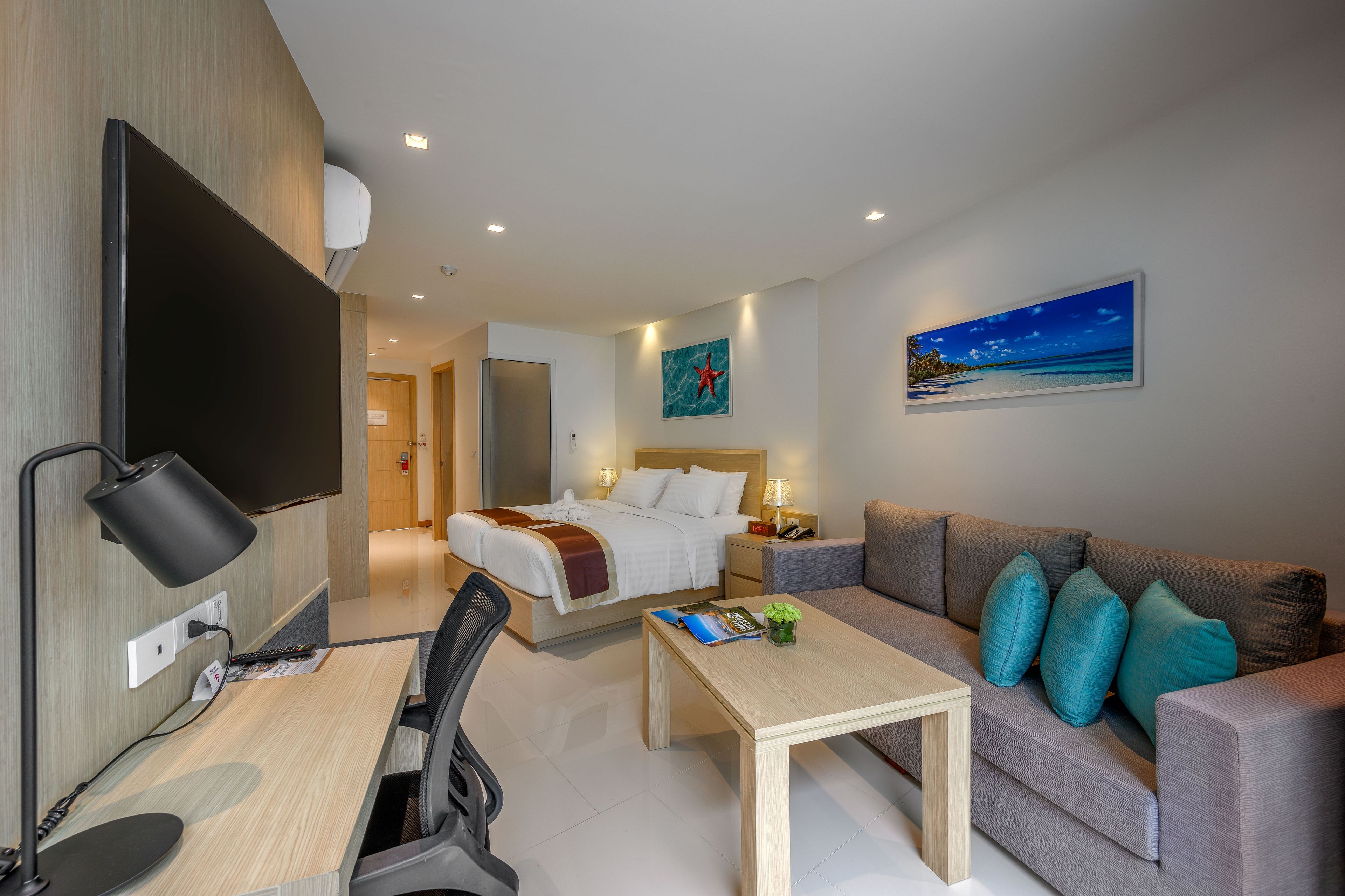 Quarto de hotel com cama, sofá, escrivaninha com computador, televisão e um quadro com uma praia.