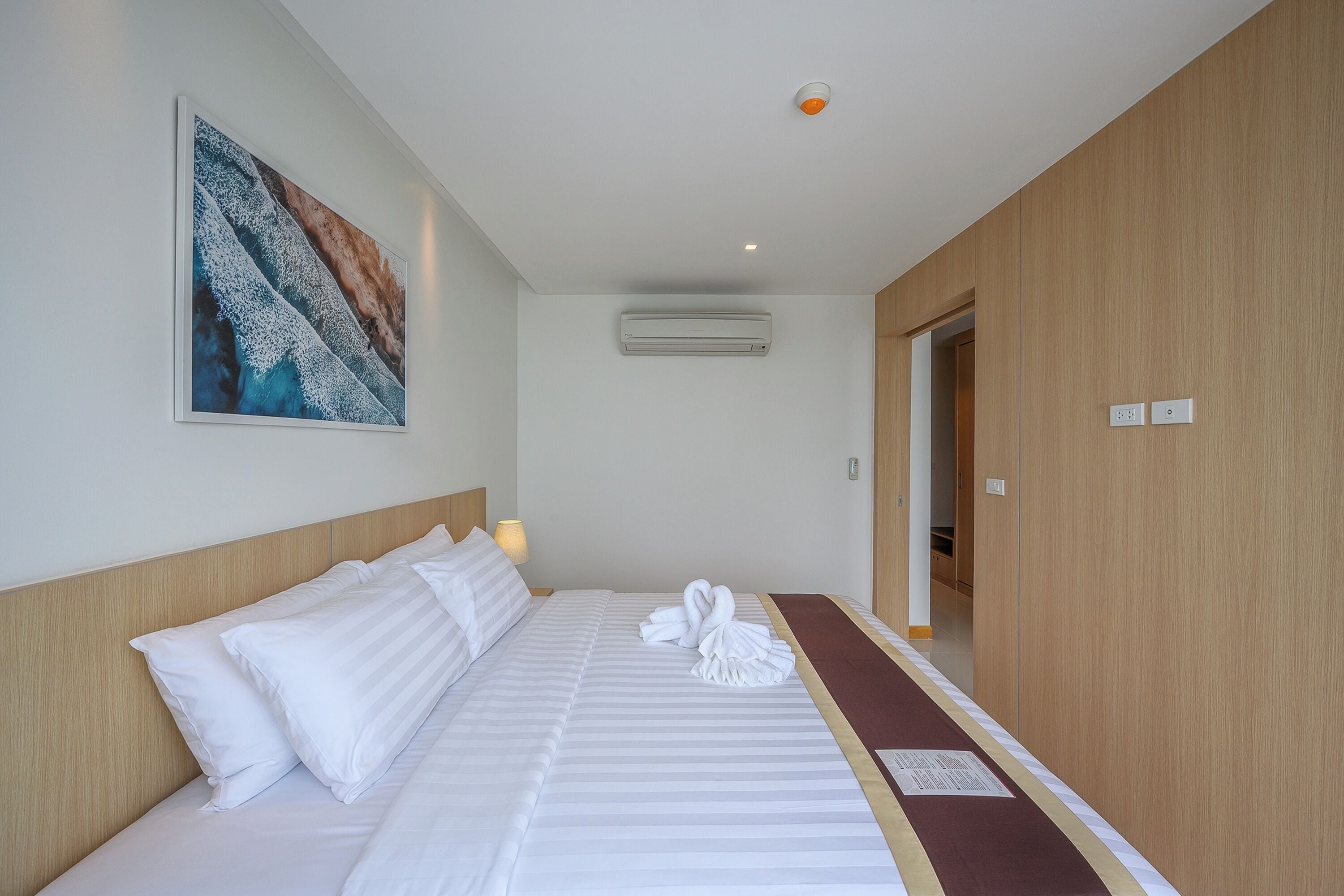Une chambre d’hôtel avec un lit soigneusement fait, un tableau accroché au mur, un appareil de climatisation et une armoire en bois.