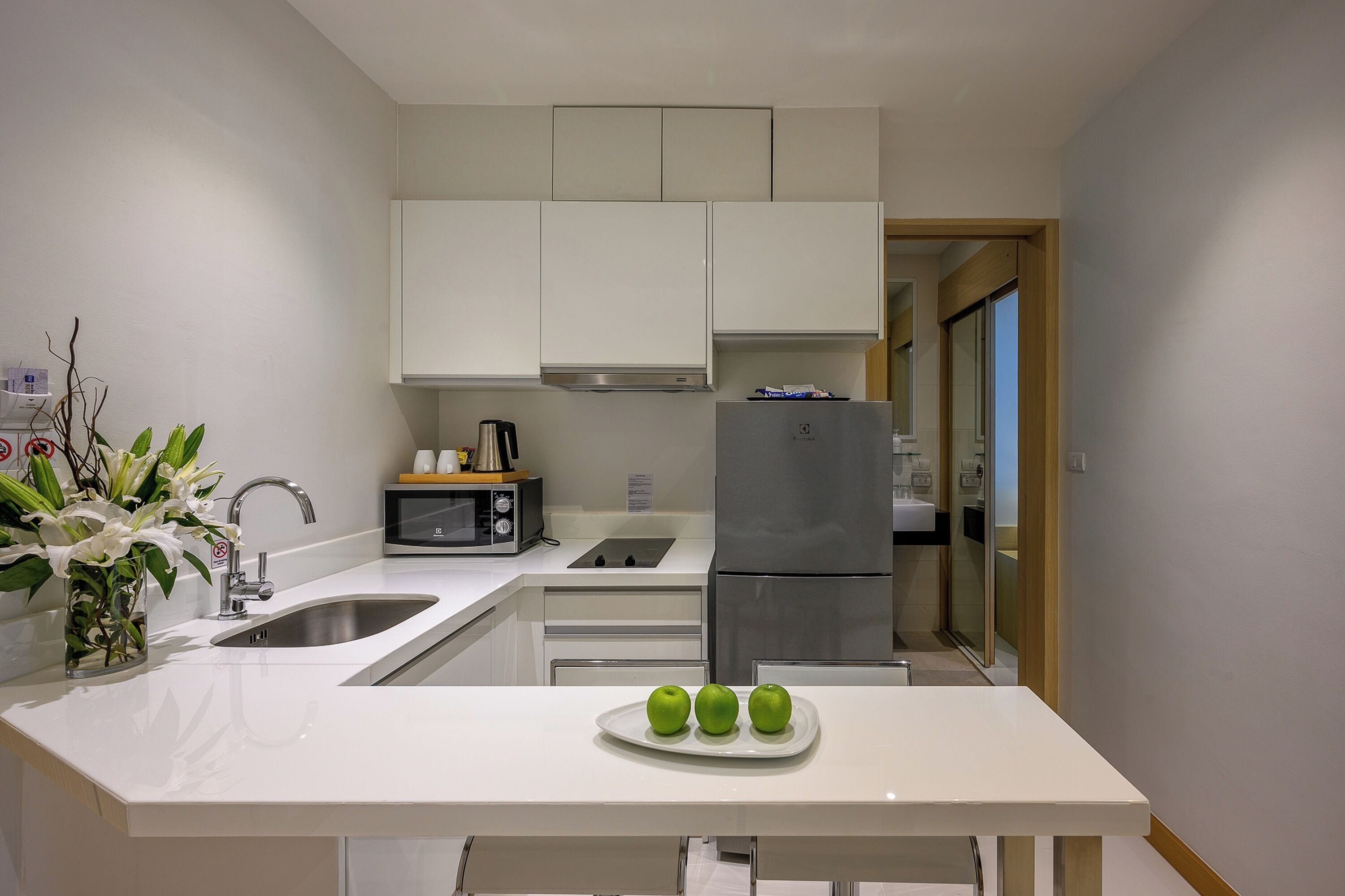 A compact kitchen with white cabinets, a stainless steel refrigerator, a microwave, and a sink. There are three green apples on a white plate on the counter.