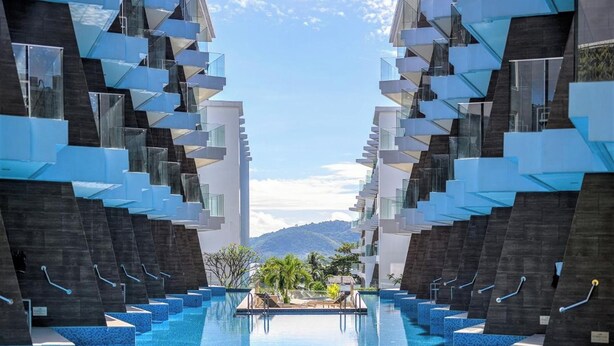 A modern hotel with a central pool, multiple balconies, and a mountainous backdrop. - The Beachfront Hotel Phuket (Rawai)