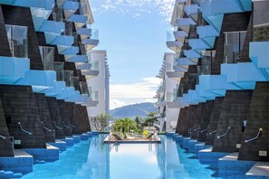 Un hotel moderno con piscina central, varios balcones y un paisaje montañoso de fondo.