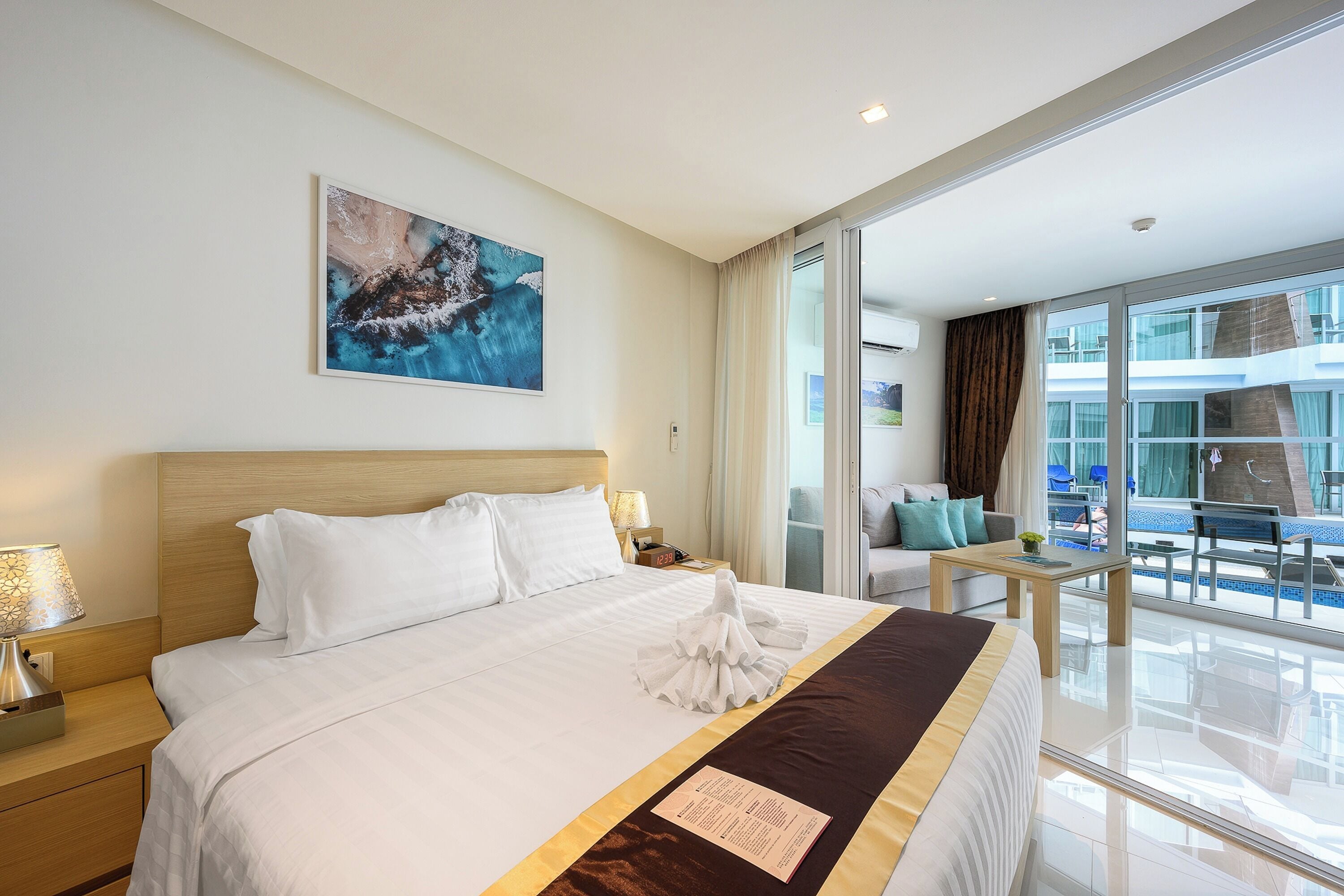 Une chambre d’hôtel avec un grand lit, un tableau au mur, un balcon donnant sur la piscine et un bureau avec une lampe.