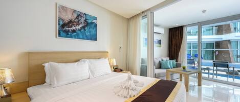 Une chambre d’hôtel avec un grand lit, un tableau au mur, un balcon donnant sur la piscine et un bureau avec une lampe.