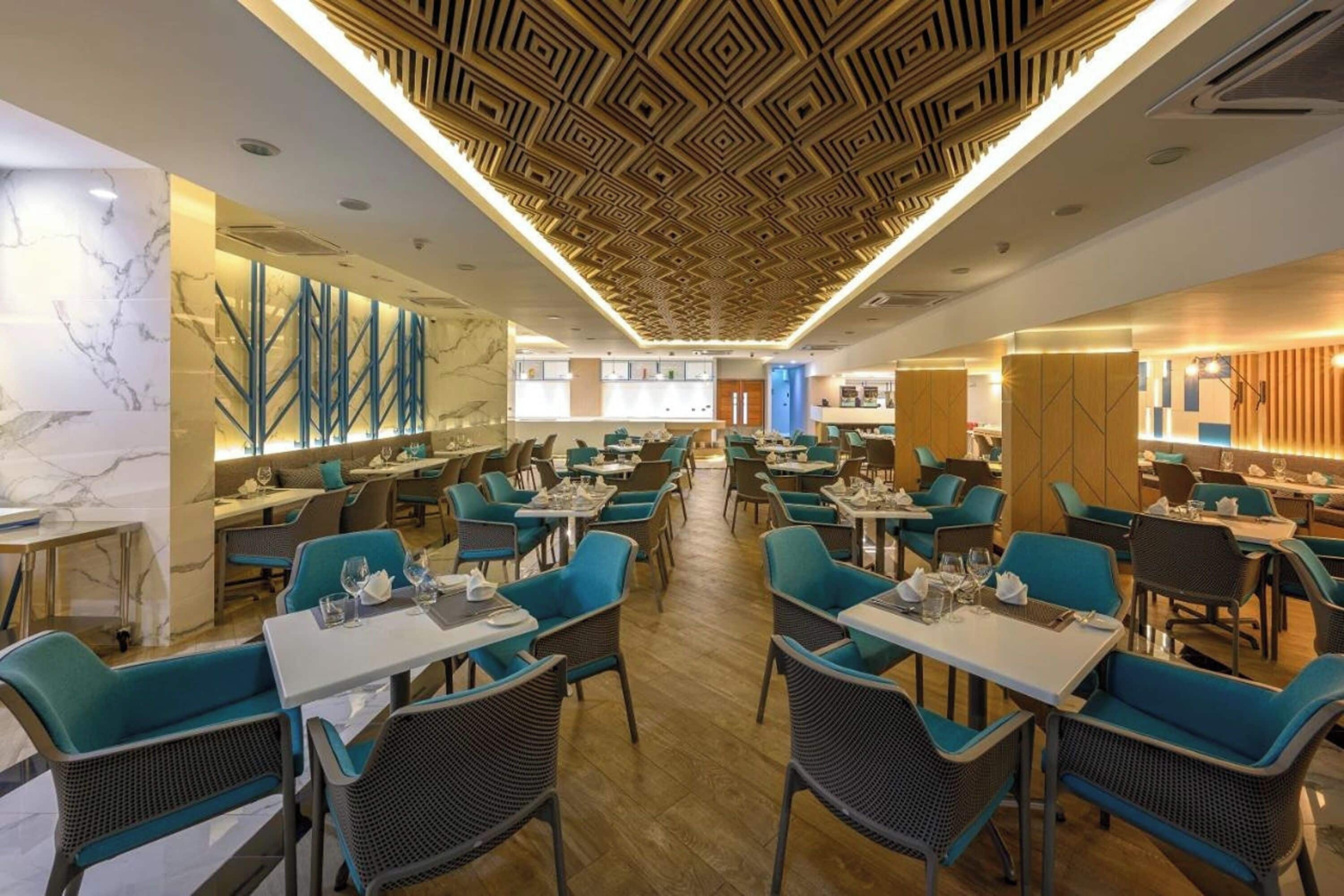 Un restaurant moderne avec des chaises recouvertes d’un tissu turquoise, des murs en marbre et un plafond orné de motifs géométriques.