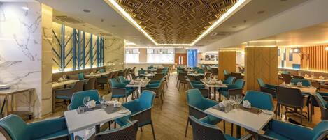 Un restaurant moderne avec des chaises recouvertes d’un tissu turquoise, des murs en marbre et un plafond orné de motifs géométriques.