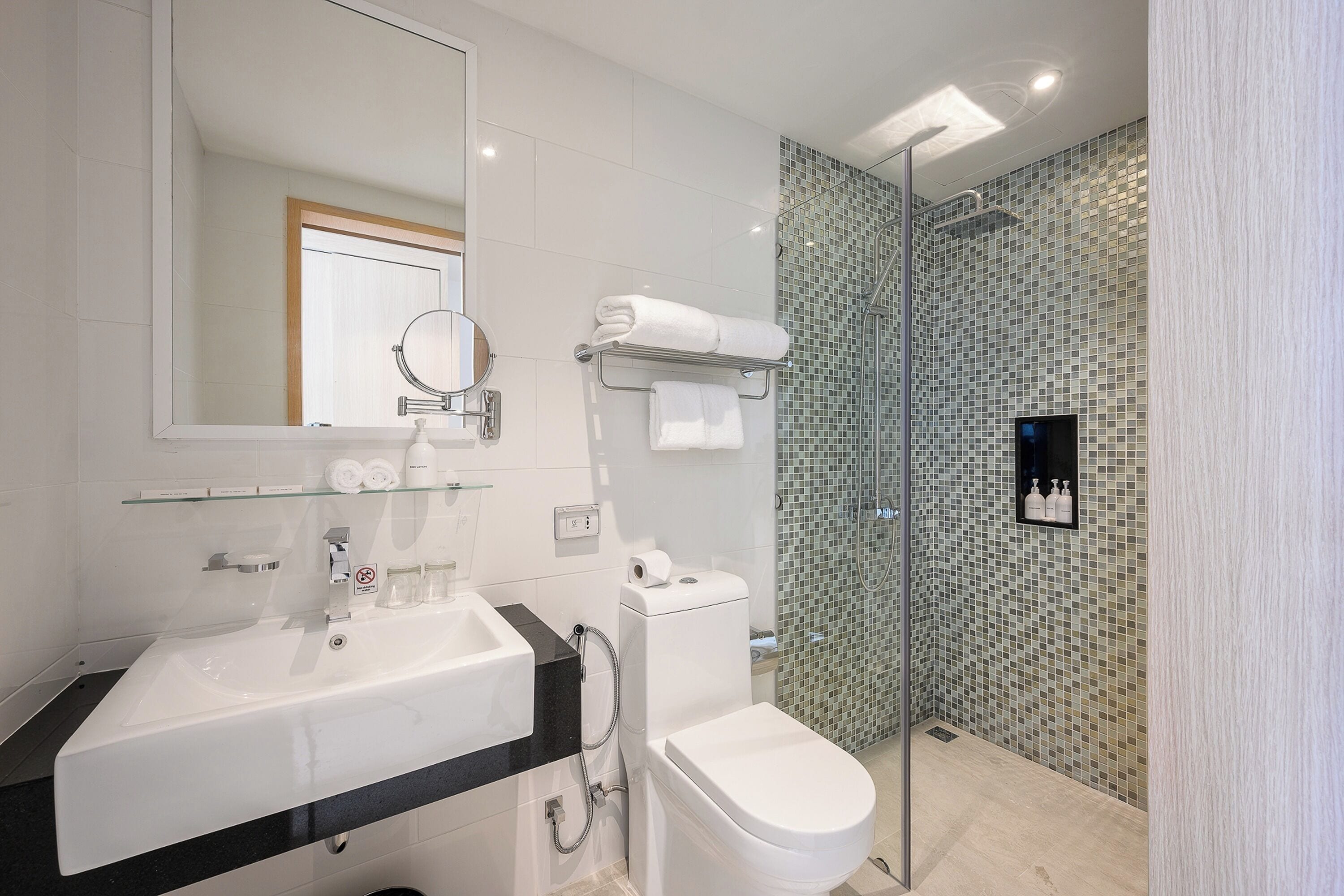 A modern bathroom with a white sink, mirror, and toilet, a glass shower enclosure, and a tiled shower wall.