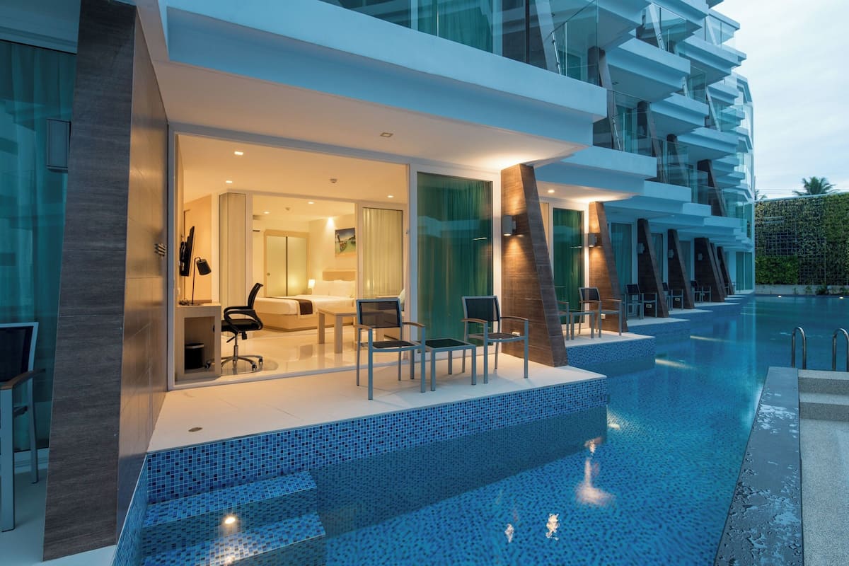 A modern hotel with a pool area, featuring a glass-enclosed room with a sofa and chairs, and a staircase leading to a balcony.