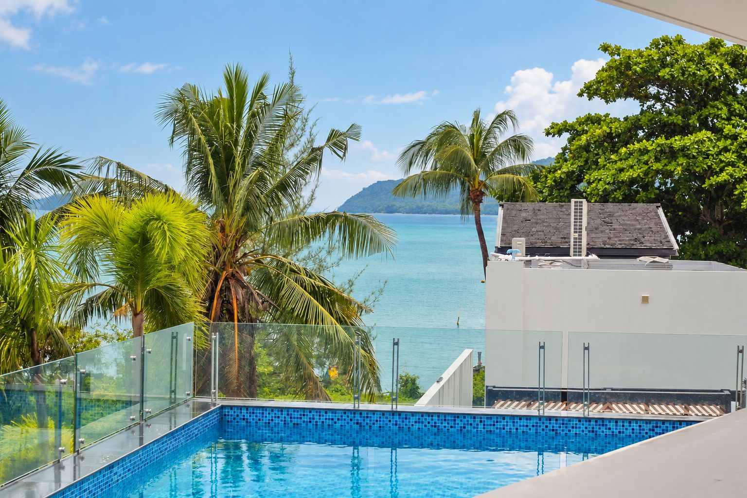 A clear blue pool with a glass barrier, surrounded by palm trees and a view of the sea.