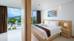 A modern hotel room with a large bed, a nightstand, a view of the sea, and a painting on the wall. - The Beachfront Hotel Phuket (Rawai)
