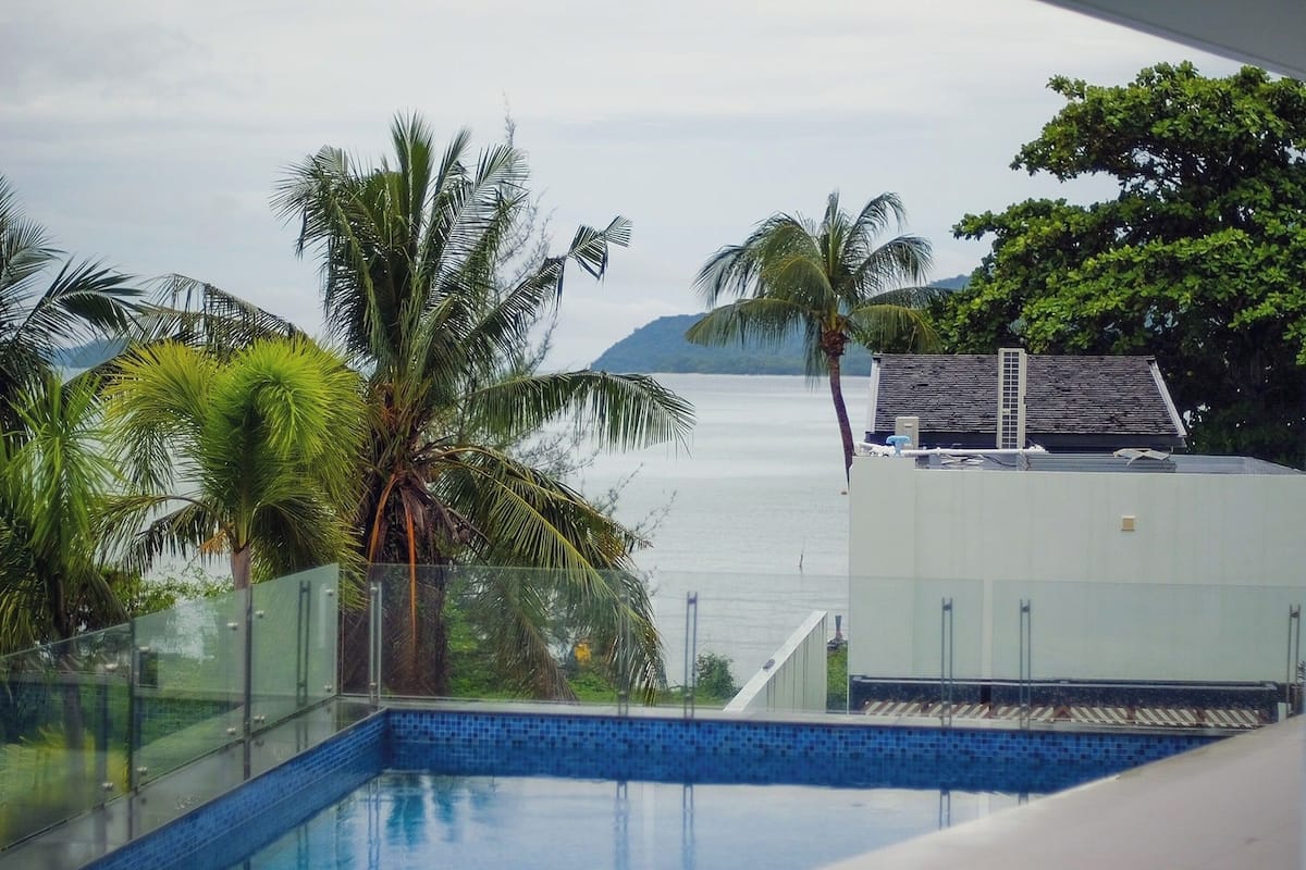 A pool with a glass railing, surrounded by palm trees and a view of the sea.