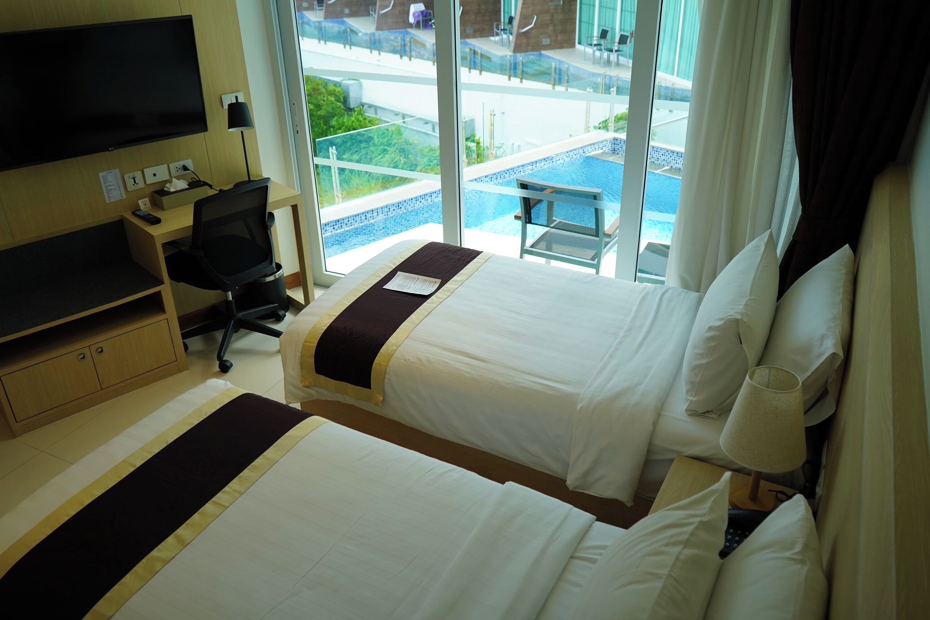 Habitación de hotel con dos camas, un escritorio con computadora, televisión y vista a la piscina y área exterior.