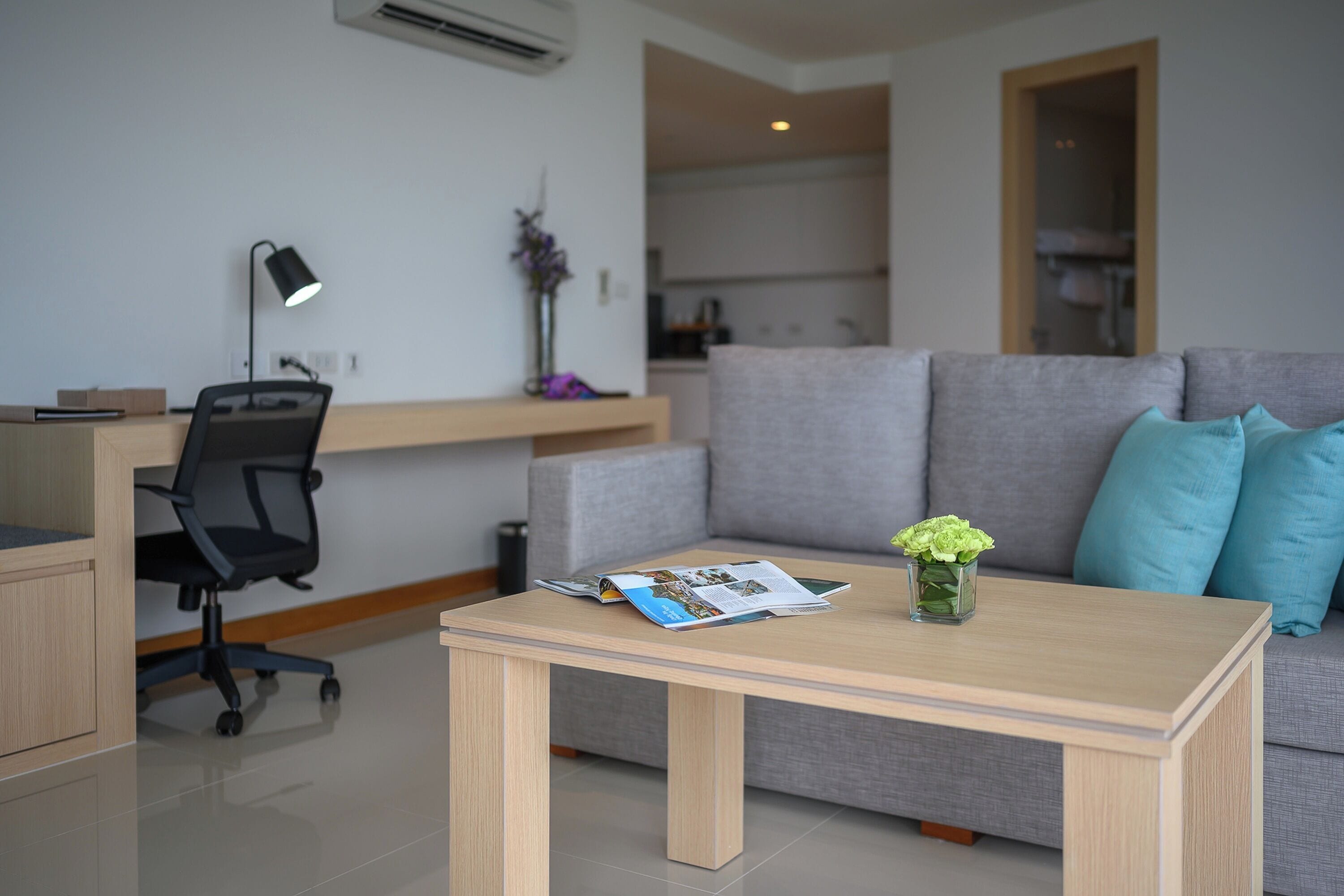 A modern living room with a grey sofa, a wooden coffee table, a black office chair, a desk with a lamp, and a vase with flowers.