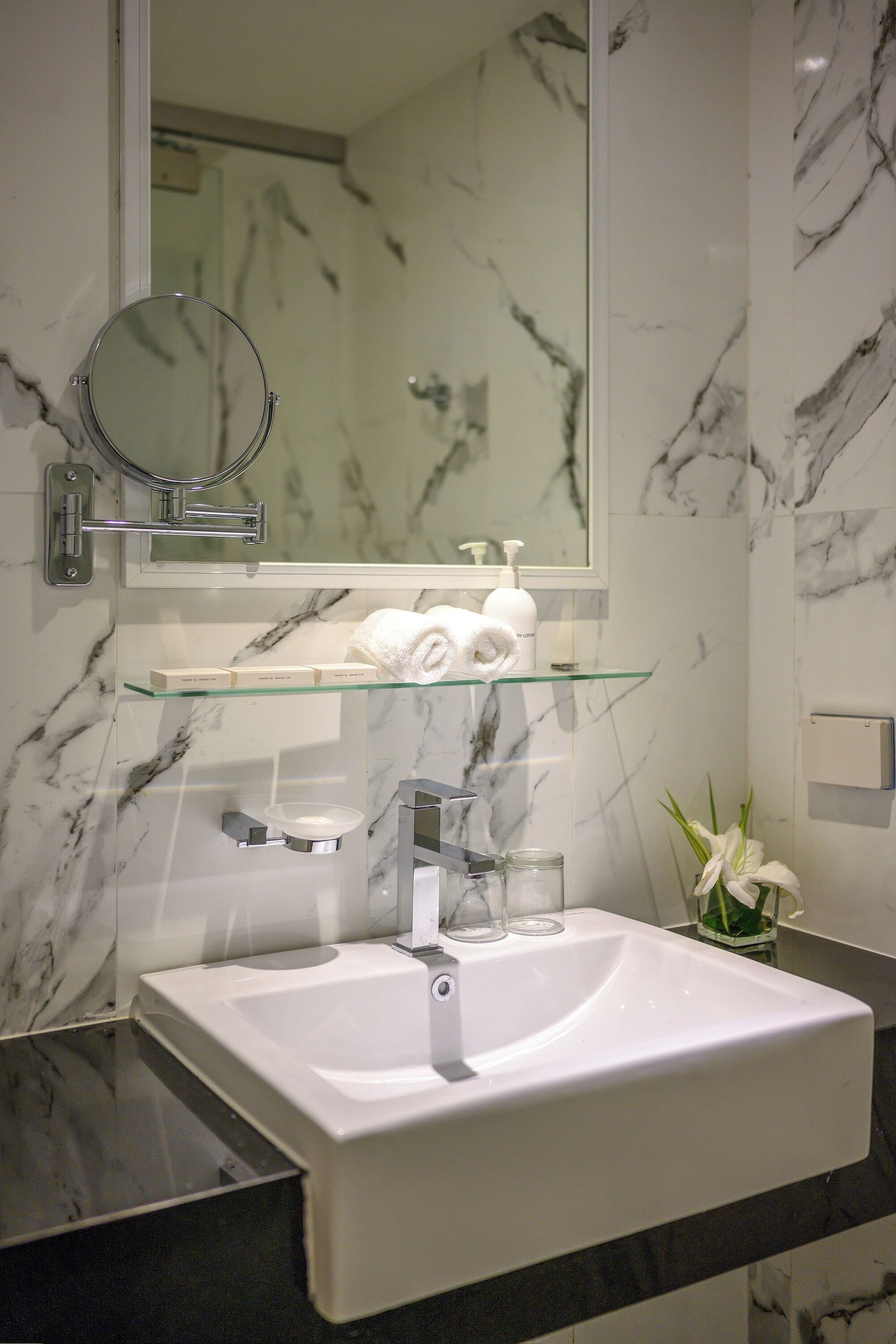 A bathroom with a white sink, a glass shelf with toiletries, a round mirror, and a marble wall.